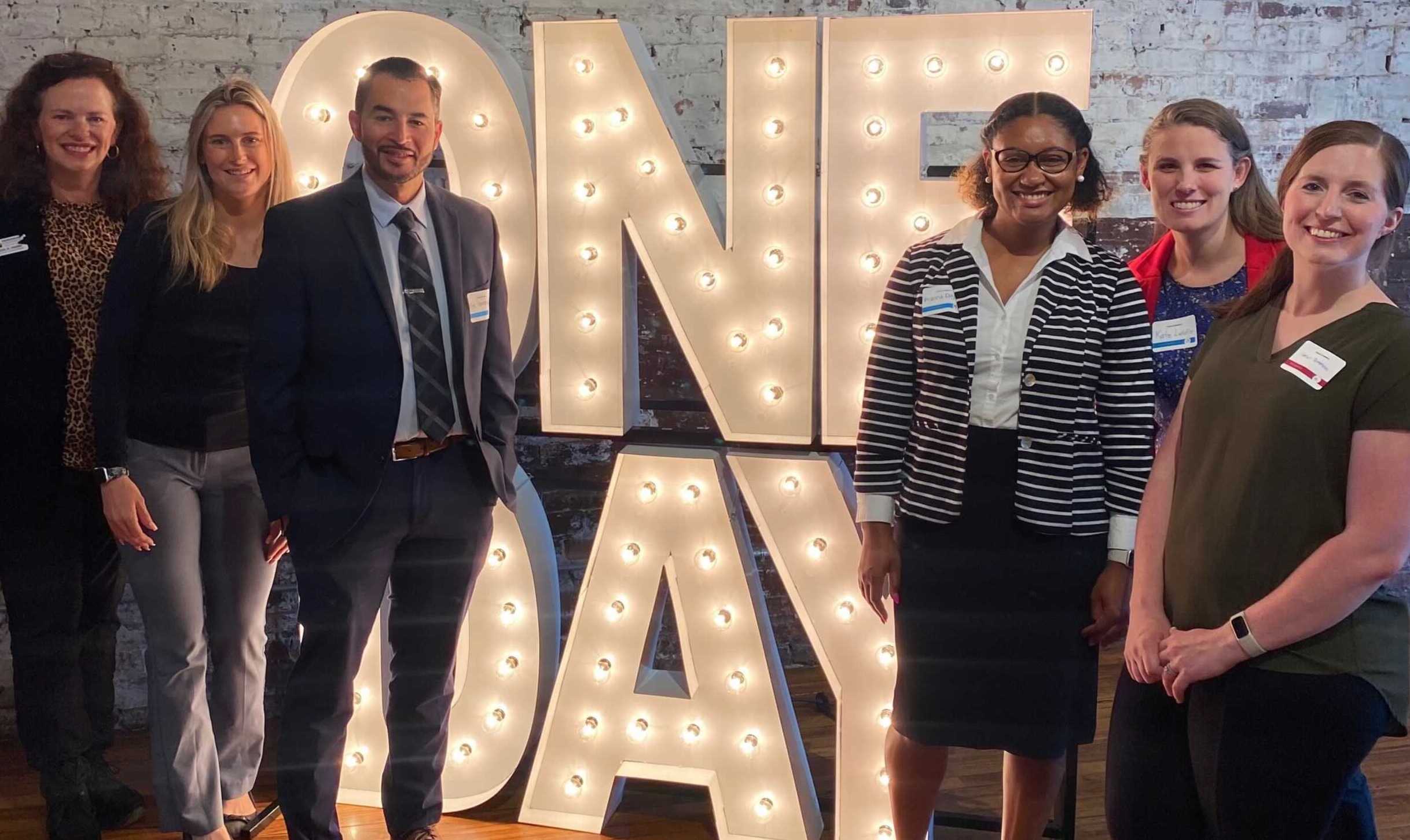 Four Teach For America alumni stand next to a One Day display
