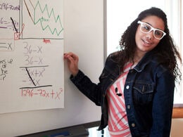 A brown-skinned, teenage female student solves a math equation on a whiteboard.