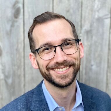Portrait of Ryan Pavel wearing glasses, light blue shirt and dark blue jacket.