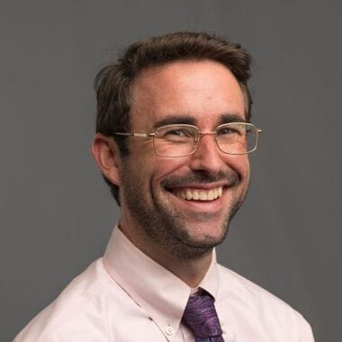 man smiling wearing a white shit and dark tie in front of a grey background