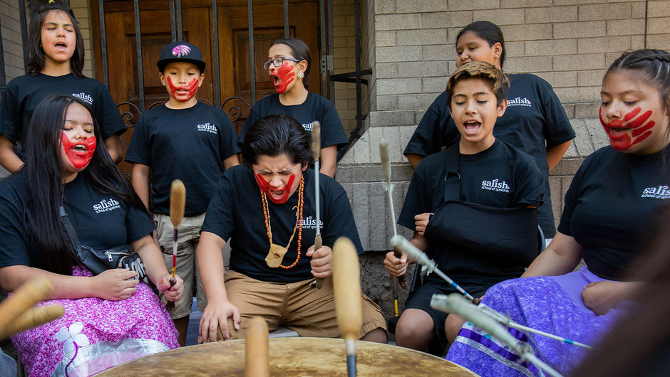 A group of students with black shirts and handprints painted over their faces in red hit a large drum while singing.