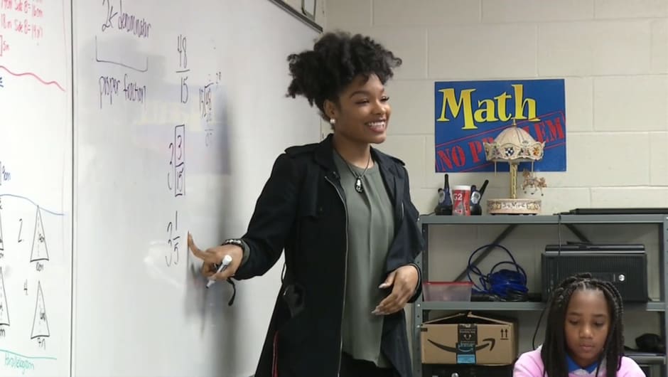 Smiling woman standing in front of whiteboard and students, pointing to math problem