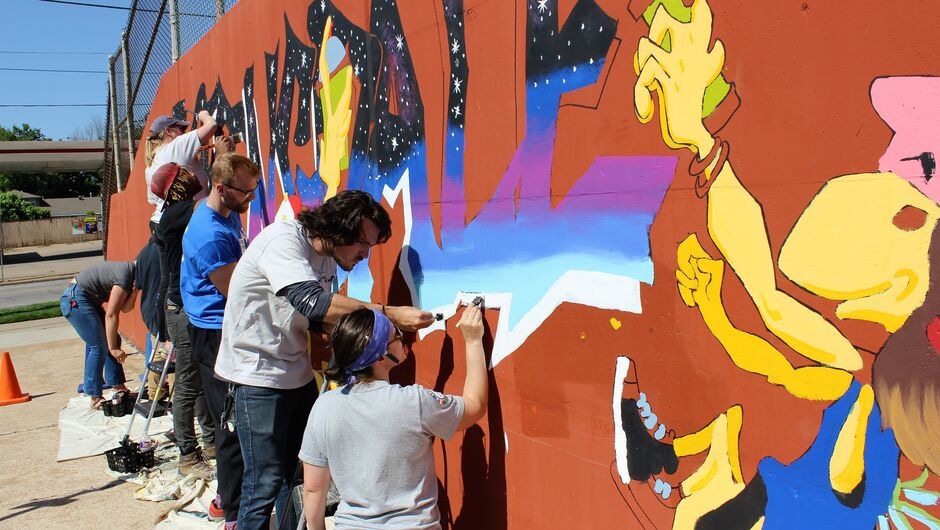 A group of individuals work on painting a colorful mural outside on a cement wall.