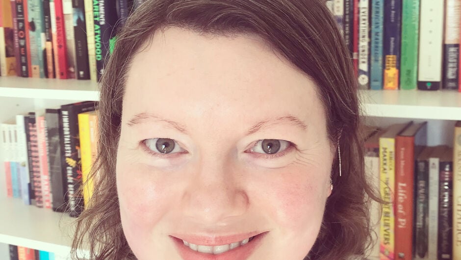 Headshot of Kate Blanchard standing in front of white bookshelves full of various books. Kate has short hair and wearing a brown sweater.