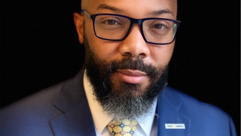 A man with a beard wearing glasses, a navy suit jacket, and a yellow tie.