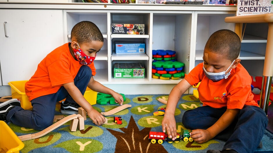 Two young Black boys in orange shirts playing with toys on a mat in a classroom.