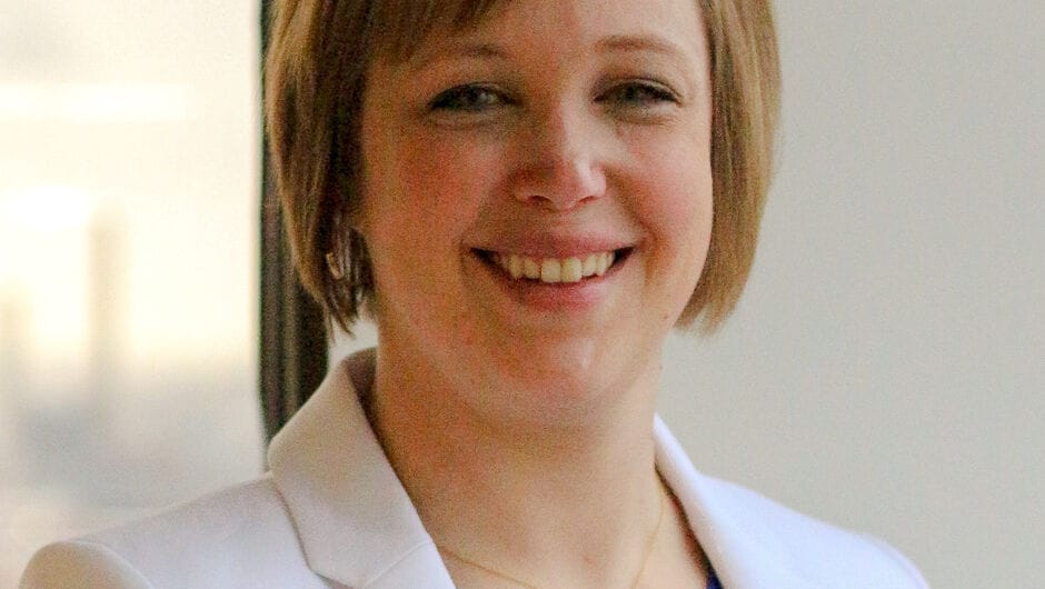 A woman with short, blonde hair wearing a blue top with a white suit jacket.