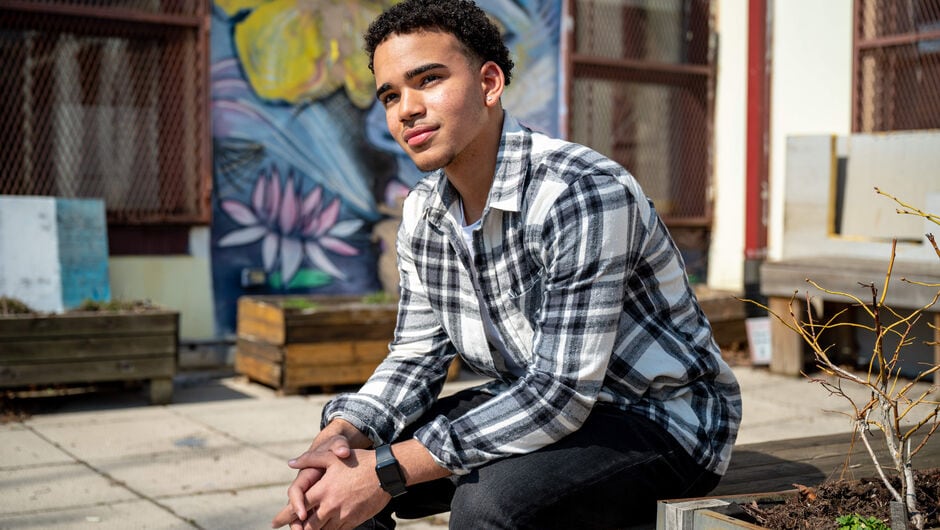 Student Juan Grullon wears a flannel shirt and sits in a courtyard with a mural in the background
