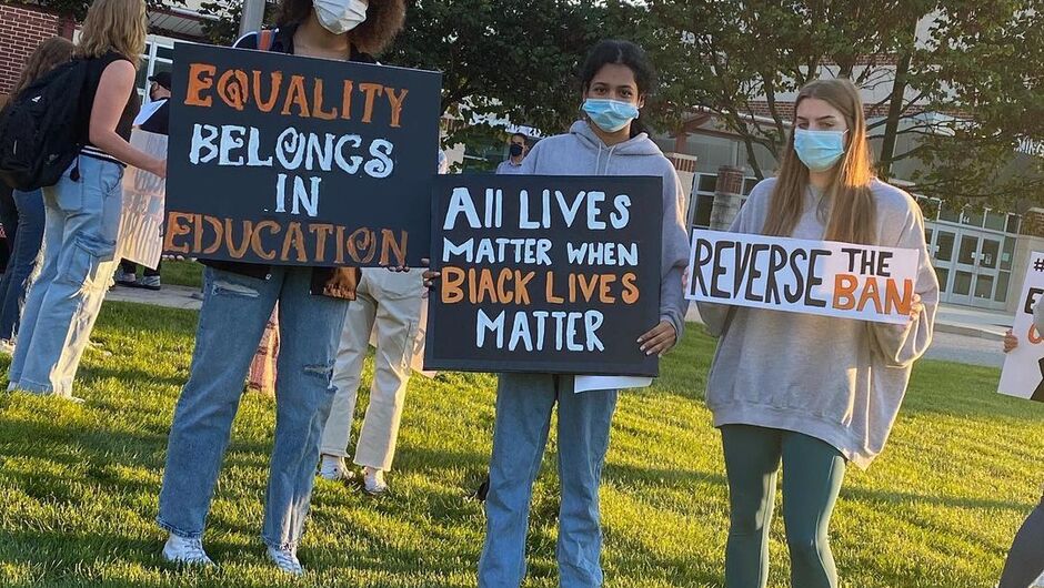 A group of students stand holding signs that say "EQUALITY BELONGS IN EDUCATION," "ALL LIVES MATTER WHEN BLACK LIVES MATTER," and "REVERSE THE BAN"