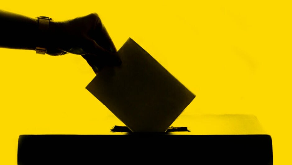 Black silhouette image in front of yellow background of a hand dropping a vote into a ballot