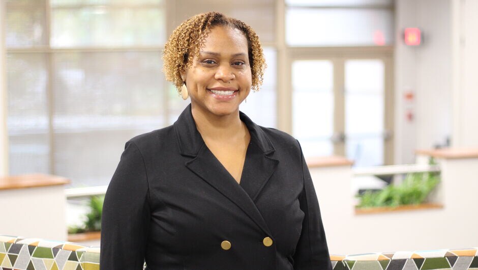 A woman in a black blazer smiles for a photo in a classroom.