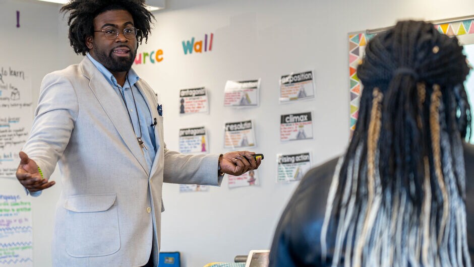 A man stands in a classroom off to the left side of the image with the back of a student's head to the right of the image