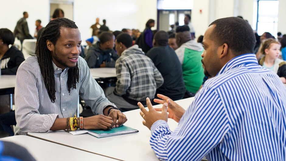 Two Black male educators face each other across a table talking. 