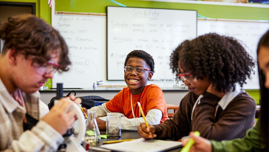 A young boy wearing glasses smiles brightly at the camera from a table in a school science lab