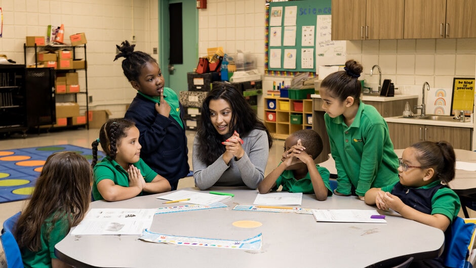 A teacher works with a small group of students at a classroom table. 