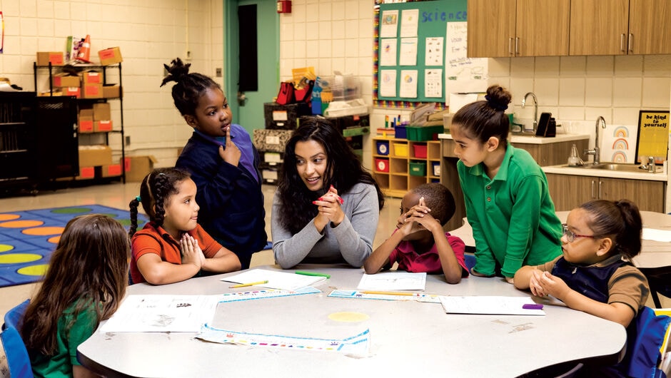 A teacher sits at a table engaging with a group of young students who gather around her.