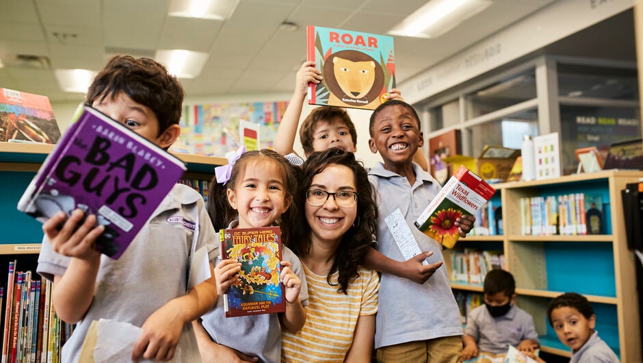 Alt text: Students excitedly hold up books with their teacher in the school library. 