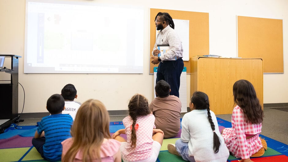A teacher leads a class of young elementary students seated on the rug.