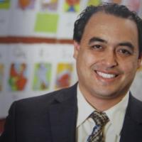 A middle-aged man with thinning black hair smiling in a classroom, wearing a black blazer, a white dress shirt, and a blue and brown patterned tie.
