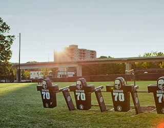 Green grassy football field, with football equipment in the foreground; a school building and blue sky in the background.