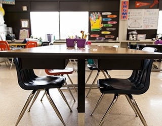 A close up photo of a student worktable with four plastic chairs inside of a classroom; other chairs and classroom materials are in the background, along with decorations on the walls and a large sunny window.