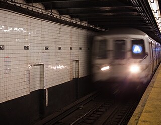 View from a subway platform of a subway train (the A train) driving away from the platform; a large wall of older white subway tile are visible in the background,