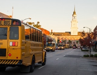 A cite street, lined with more than 5 yellow school buses; a large city hall building in visible at the end of the street; the sun in rising off-camera.