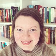 Headshot of Kate Blanchard standing in front of white bookshelves full of various books. Kate has short hair and wearing a brown sweater.