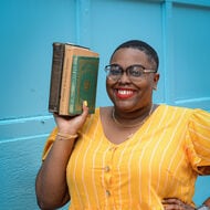 Dawnavyn James, a Black woman, in a yellow dress stands in front of a blue wall while holding books parallel to her face