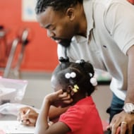 A teacher works with a young student at her desk.